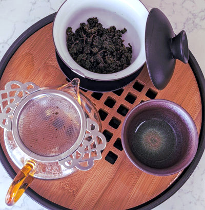 gaiwan filled with tea leaves, small teacup, and small glass pitcher all on a round tea tray