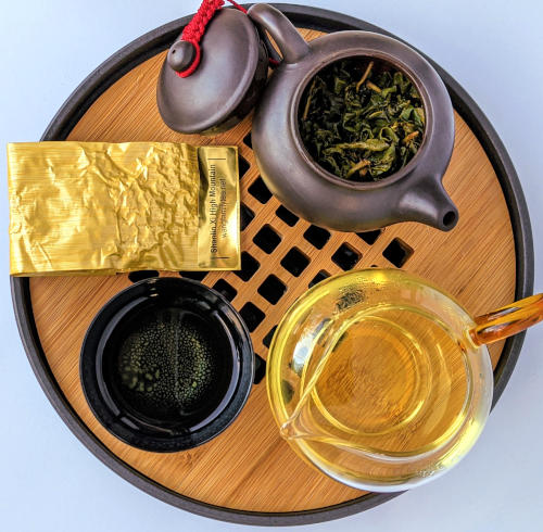 A top down image of an open yixing teapot with tea leaves inside, a small cup, a small glass pitcher filled with amber liquid, and a small golden bag, all on a round wooden tea tray.